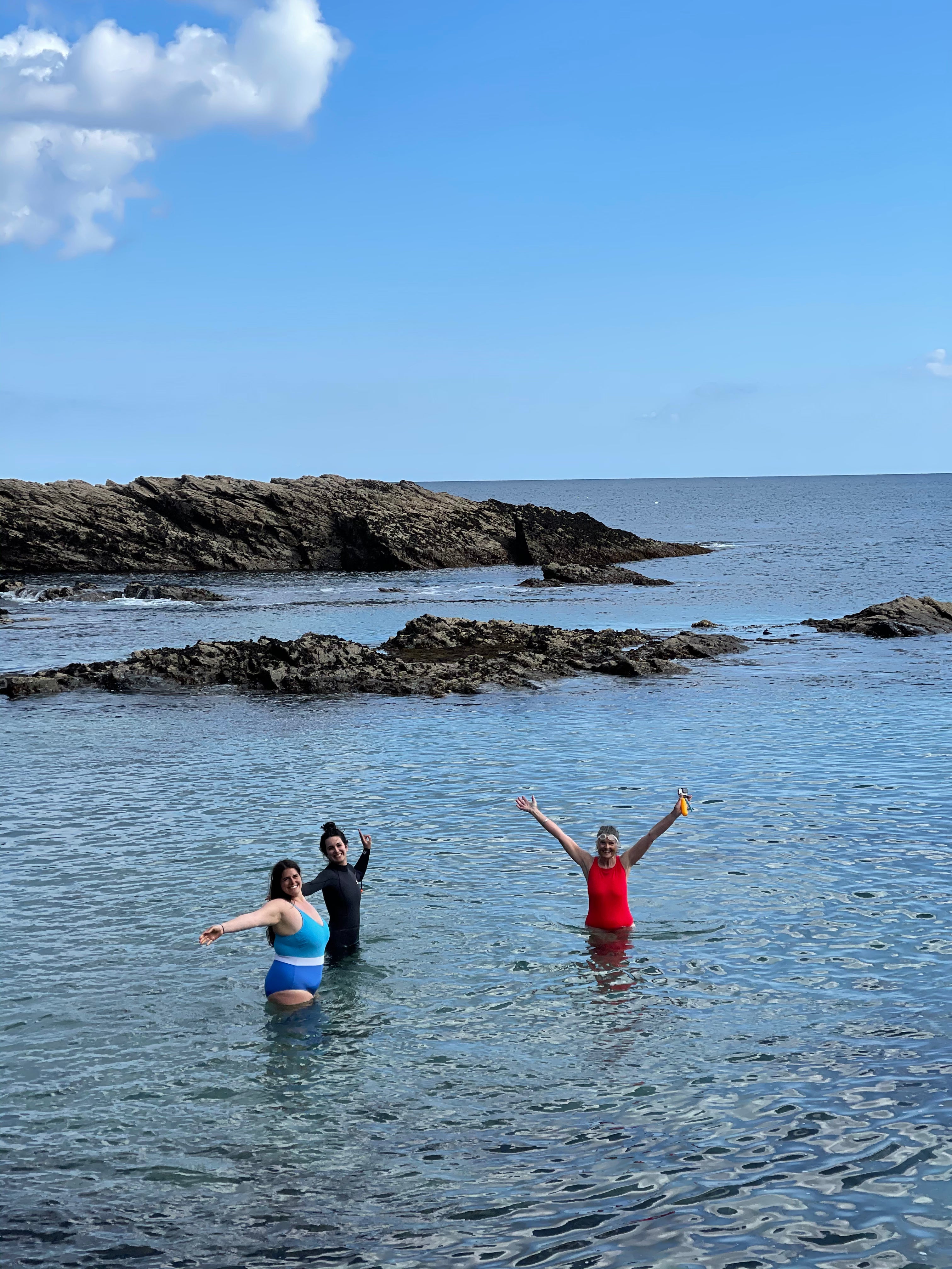 Three people standing in shallow water near rocky shores with a clear blue sky.