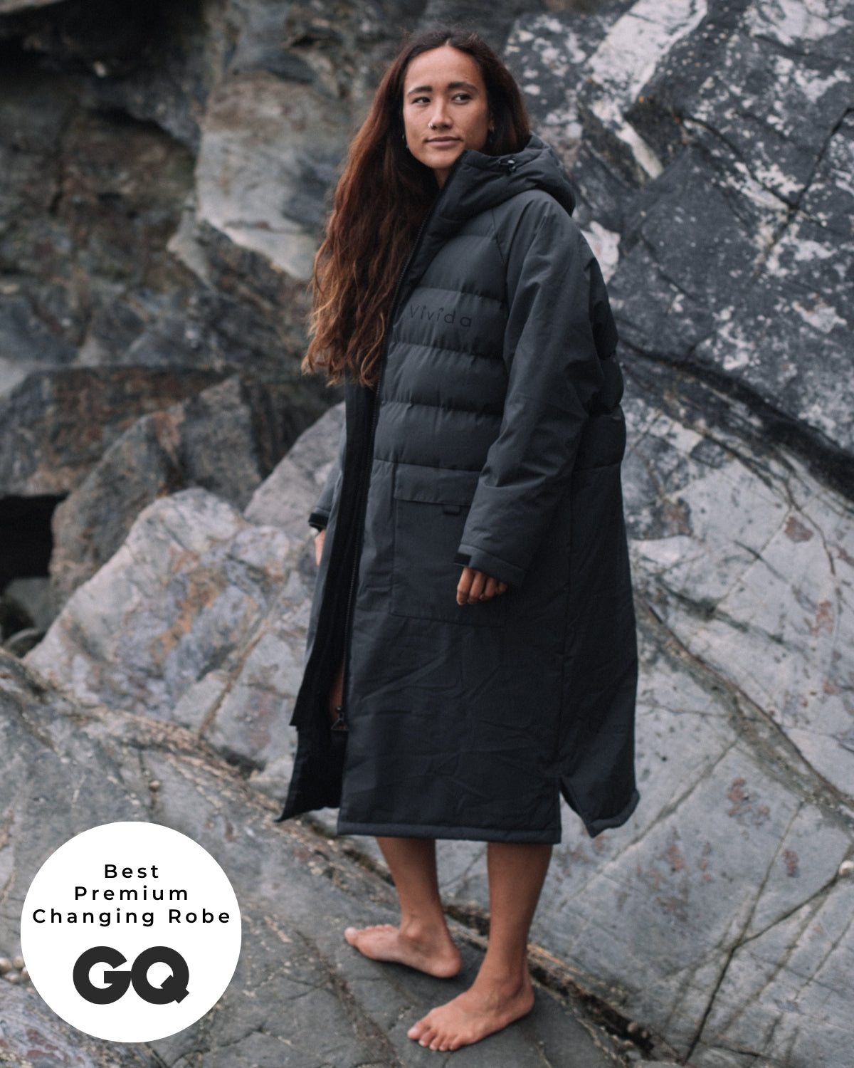 Lead_women - Woman wearing a Vivida Lifestyle All Weather Puffer Changing Robe, Fossil Grey Drying Robe for swimming standing on a rock. Sticker: Best Premium Changing Robe - GQ magazine.