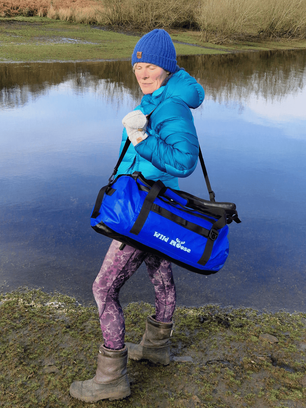 lady wild swimmer stood by water carrying an electric blue duffel bag on her shoulder
