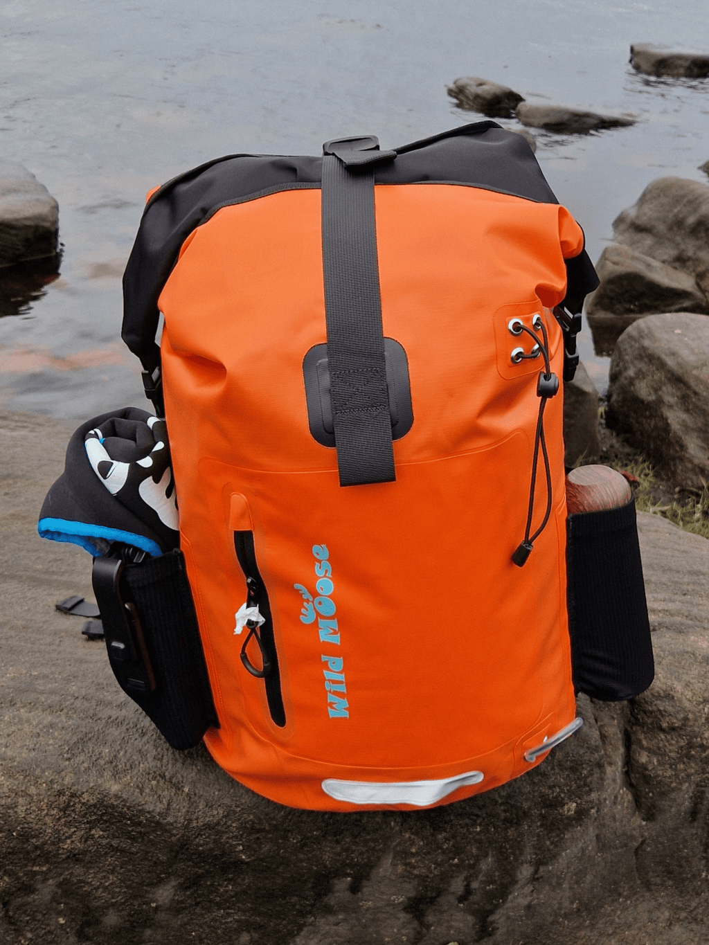 Wild Moose Bright orange waterproof backpack for open water swimming, placed on rocky shoreline
