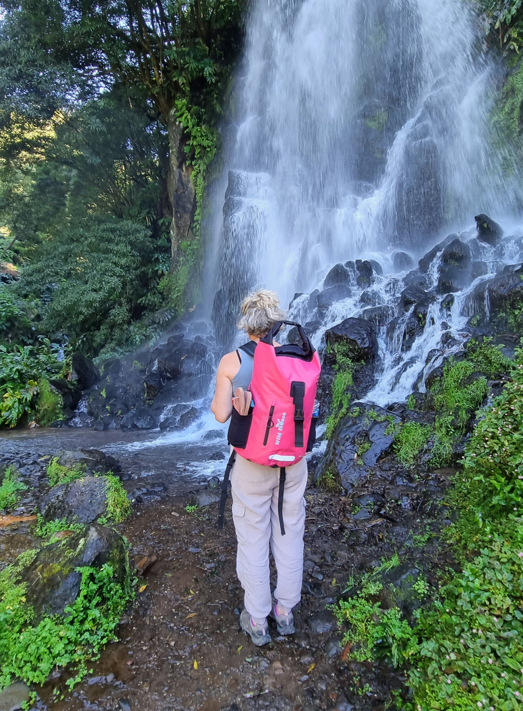 Wild Swimmer wearing bright pink waterproof swim backpack by Wild Moose, by waterfall for Cold Water Dippers.

