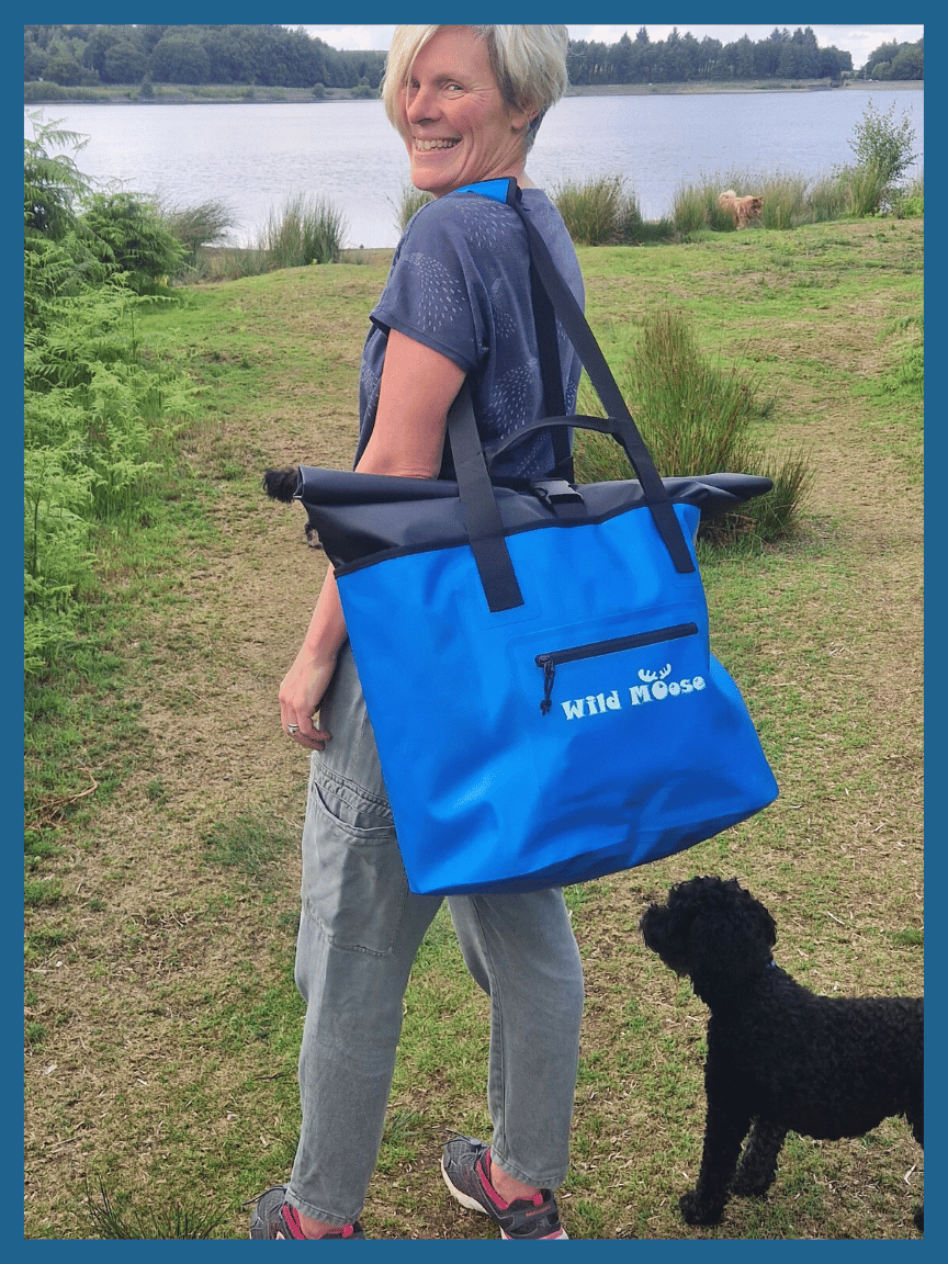 A woman with a Wild Moose Waterproof Eco Bag in Bright Blue Stood by a Lake with a Dog