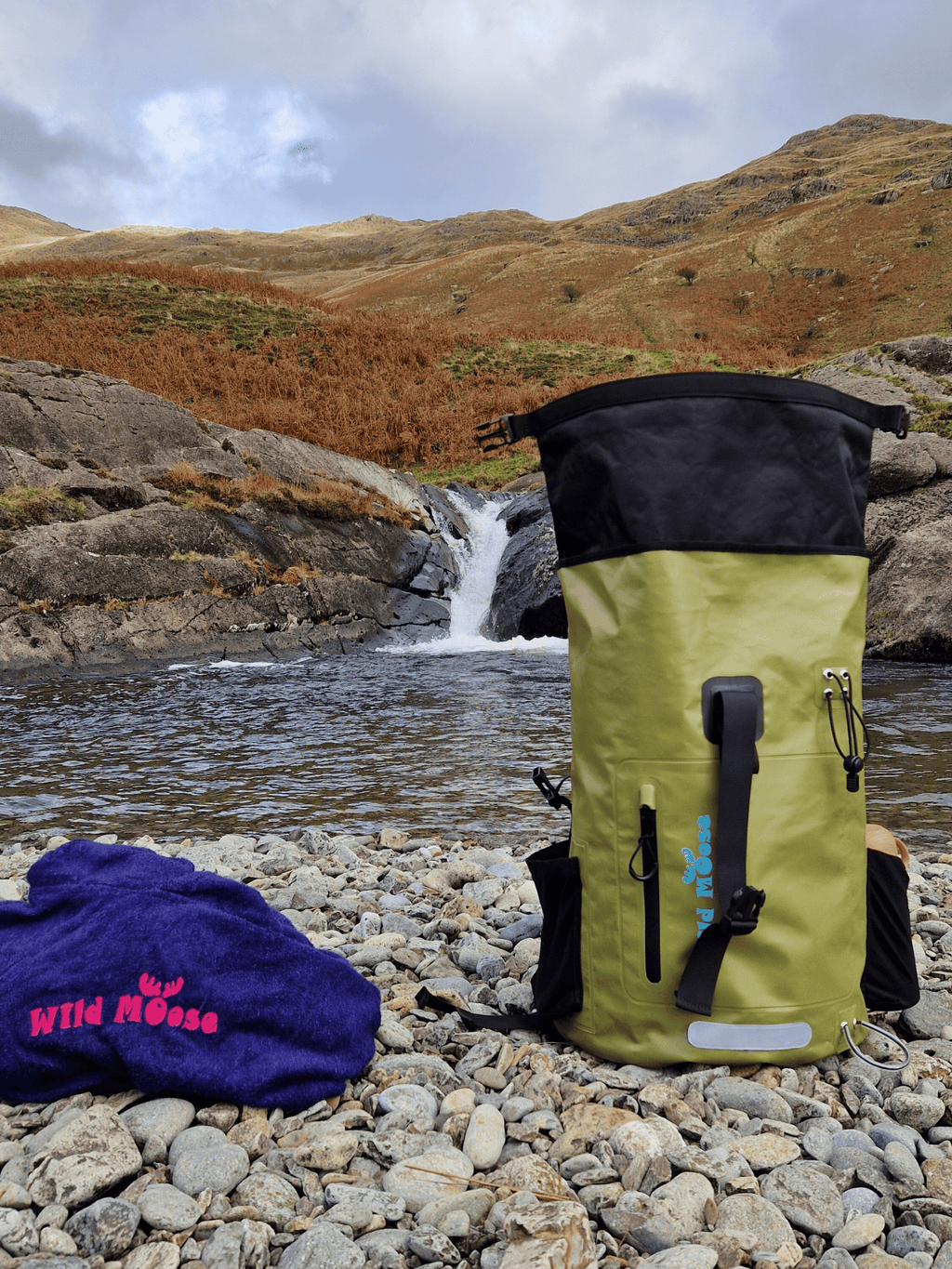 Green waterproof swim backpack by Wild Moose, placed on rocky shore beside waterfall wild swimming spot.
