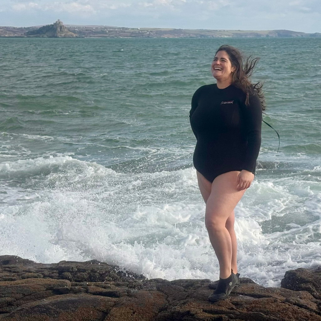Plus size woman wearing black long sleeve swimsuit, stood on rocks with St Michaels Mount swim spot - ideal for wild swimmers
