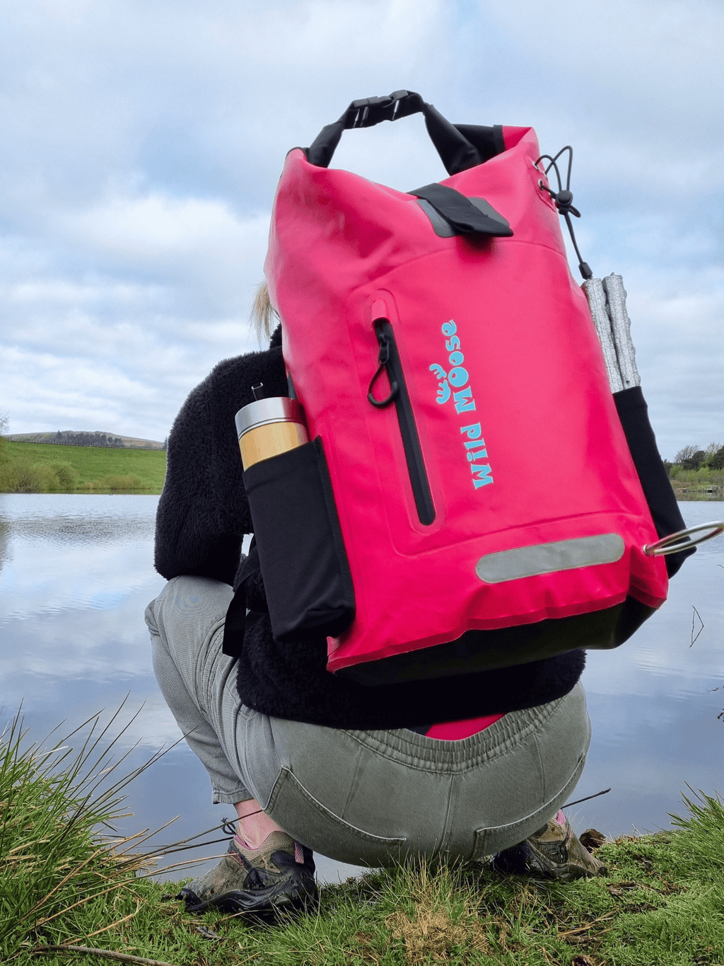 Woman with a bright pink waterproof 45L backpack by Wild Moose on her back, crouching down by a wild swimming lake, designed for wild swimmers, cold water swimmers, and open water adventurers.