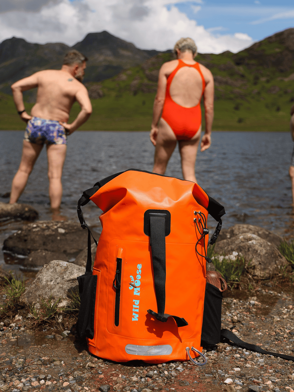 Bright orange waterproof backpack for open water swimming, placed on rocky shoreline by Swimming Lake with Wild Swimmers going for a Cold Water Dip.
