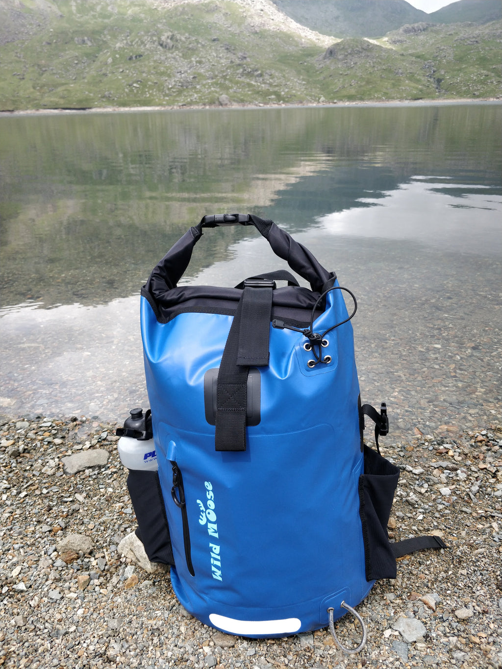 Bright blue waterproof Wild Moose backpack lakeside by a wild swimming spot.