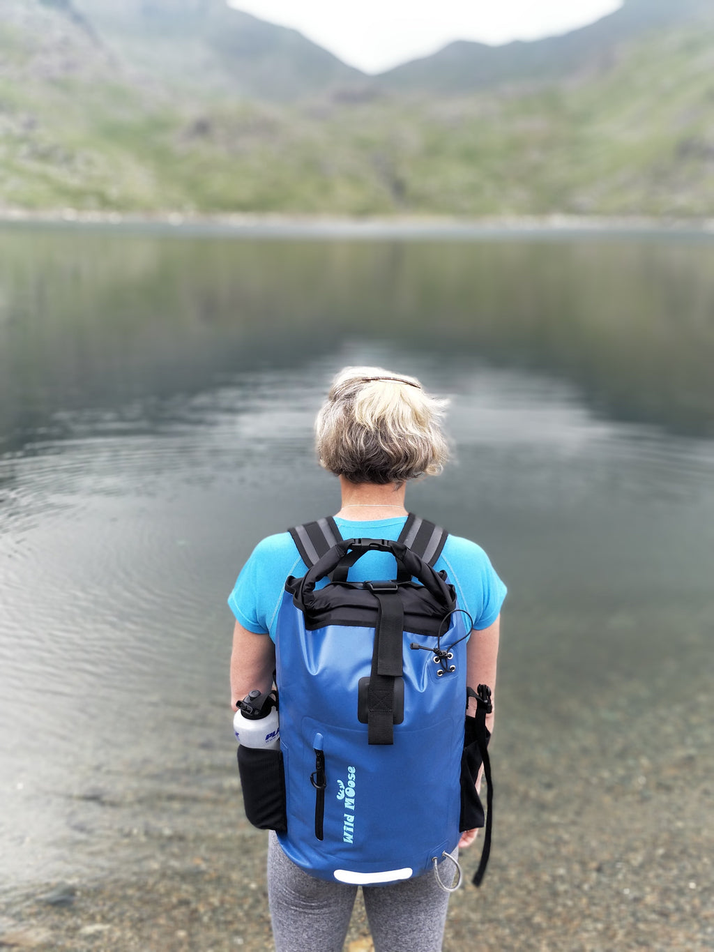 Wild Swimmer stood by wild swimming lake wearing a blur waterproof Wild Moose Backpack.