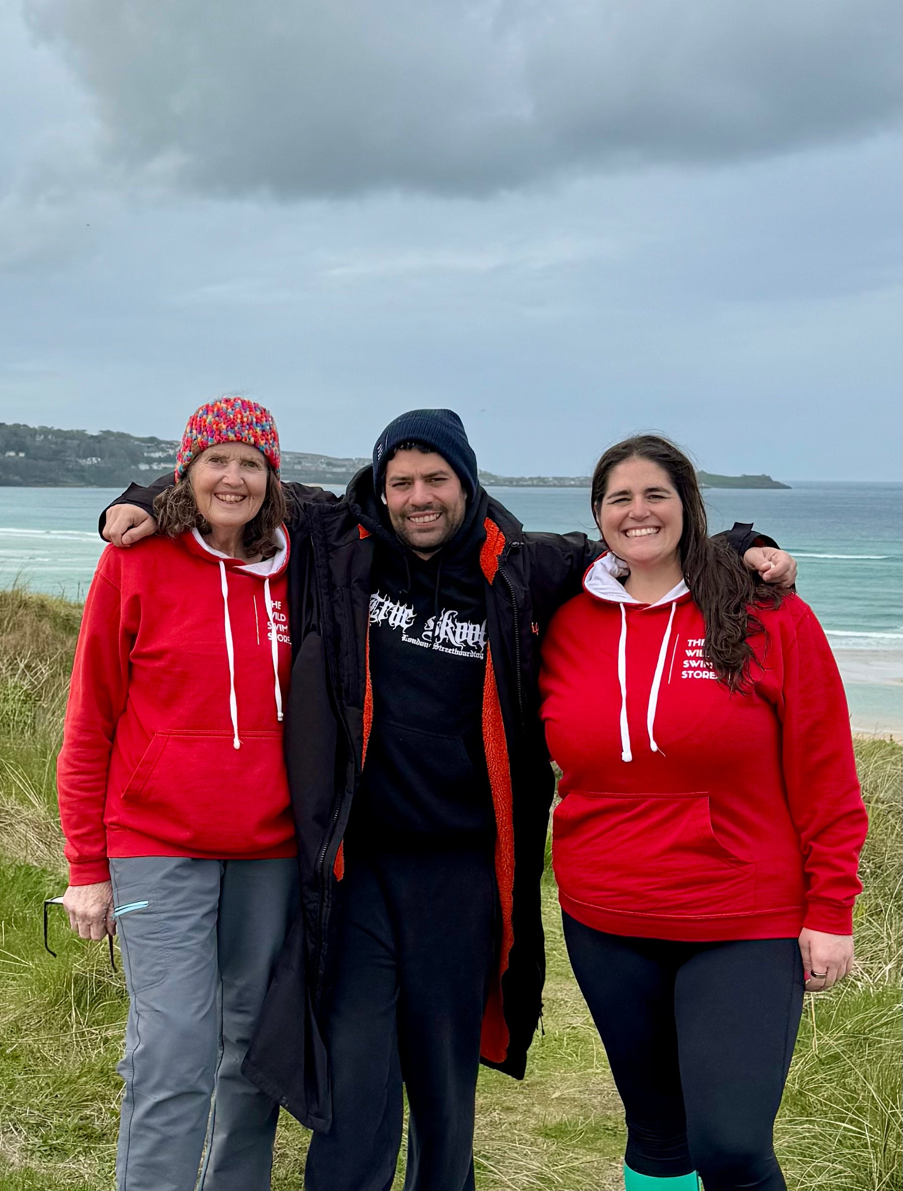 Tam, Jay and Mama Swim from The Wild Swim Store stood together wearing bright red Wild Swimming hoodie by the sea