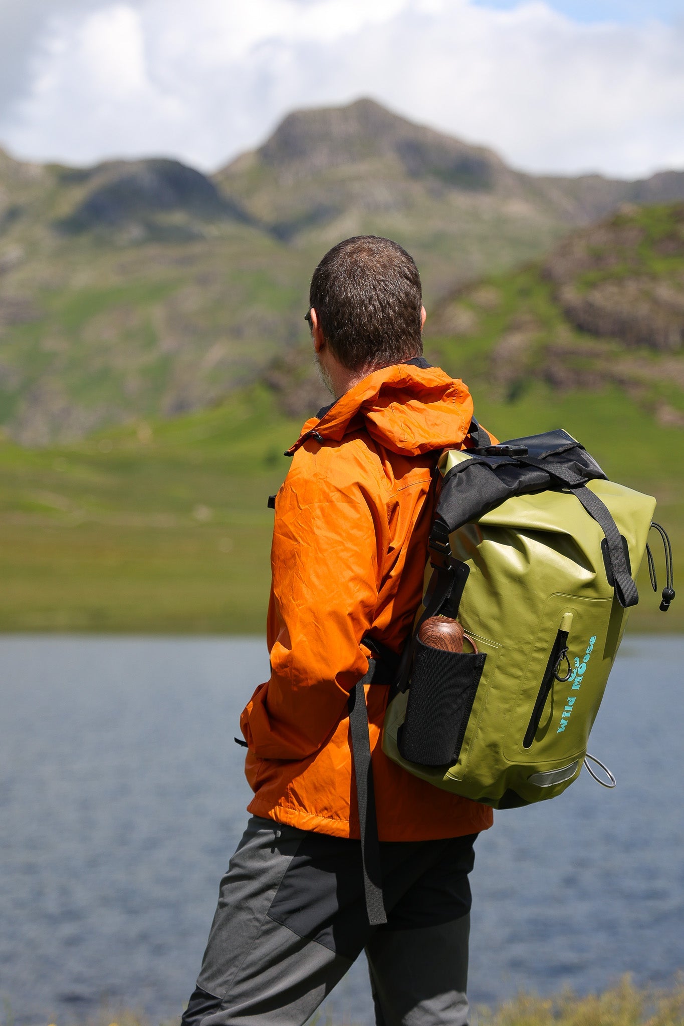 man in orange coat and carrying green backpack looking out over a tarn at a mountain view