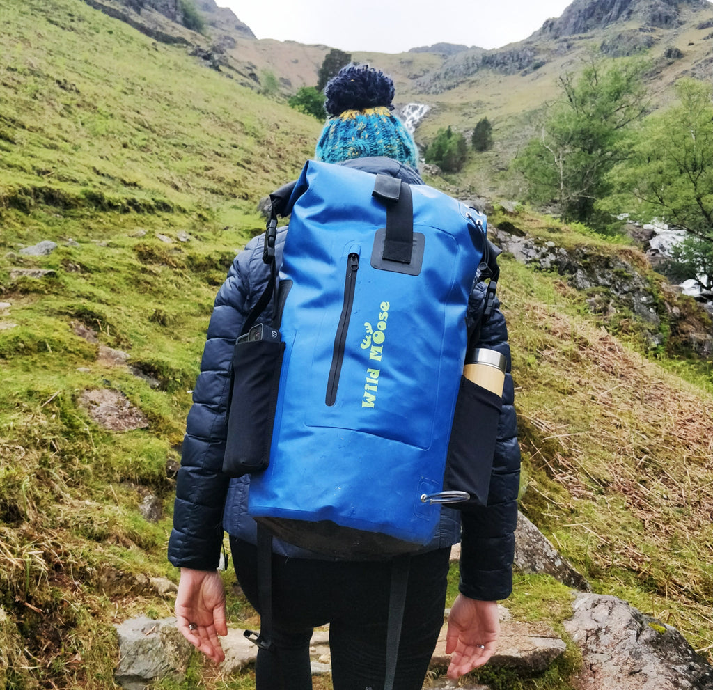 Outdoor shot of a person wearing a blue waterproof hiking backpack while ascending a rugged mountain track with walking poles, featuring a drinks bottle attached—ideal for hikers and outdoor adventurers