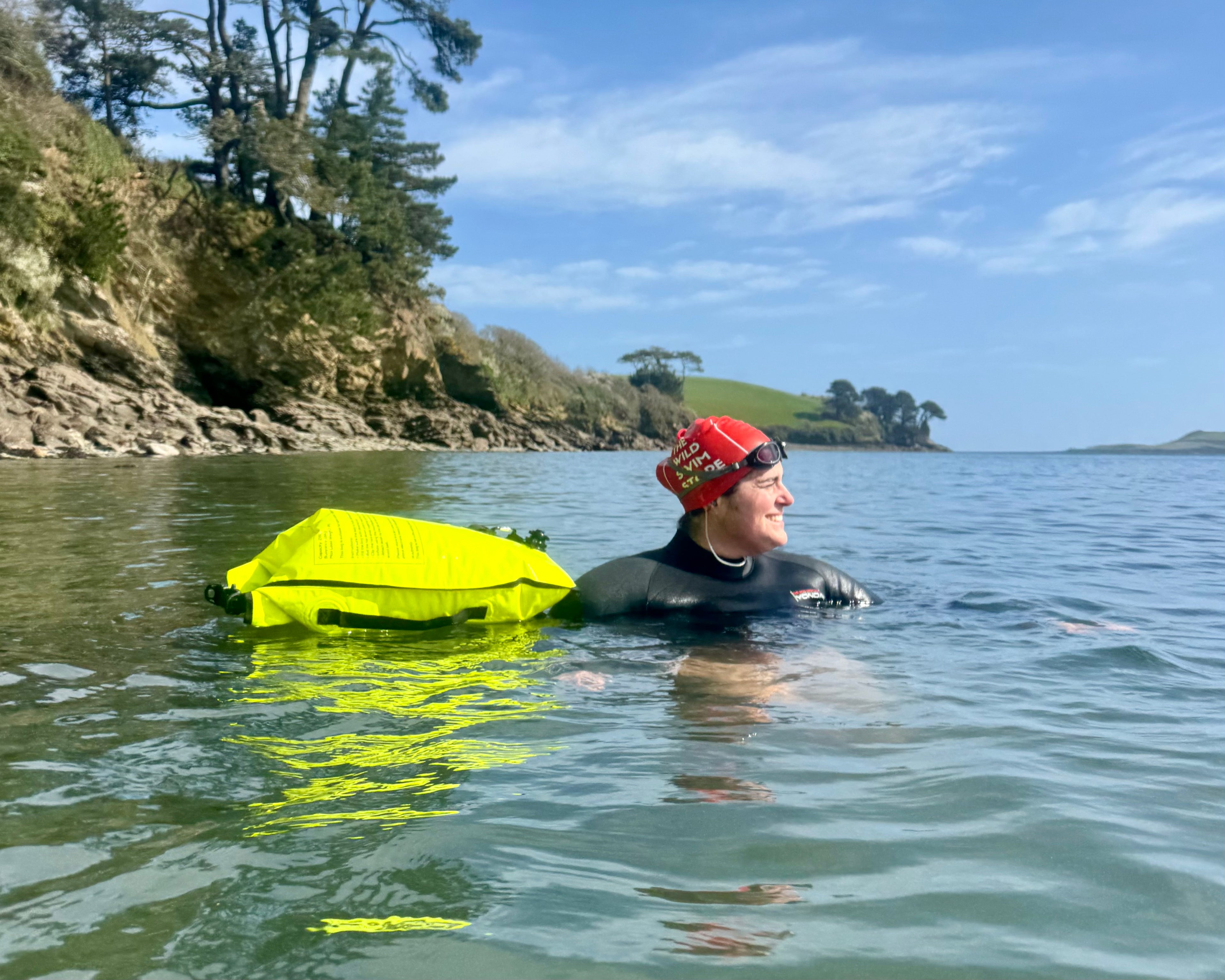 Wild swimmer wearing a wetsuit and red swim cap with goggles, floating in the sea with a bright yellow tow float, rocky coastline and blue sky in the background.
